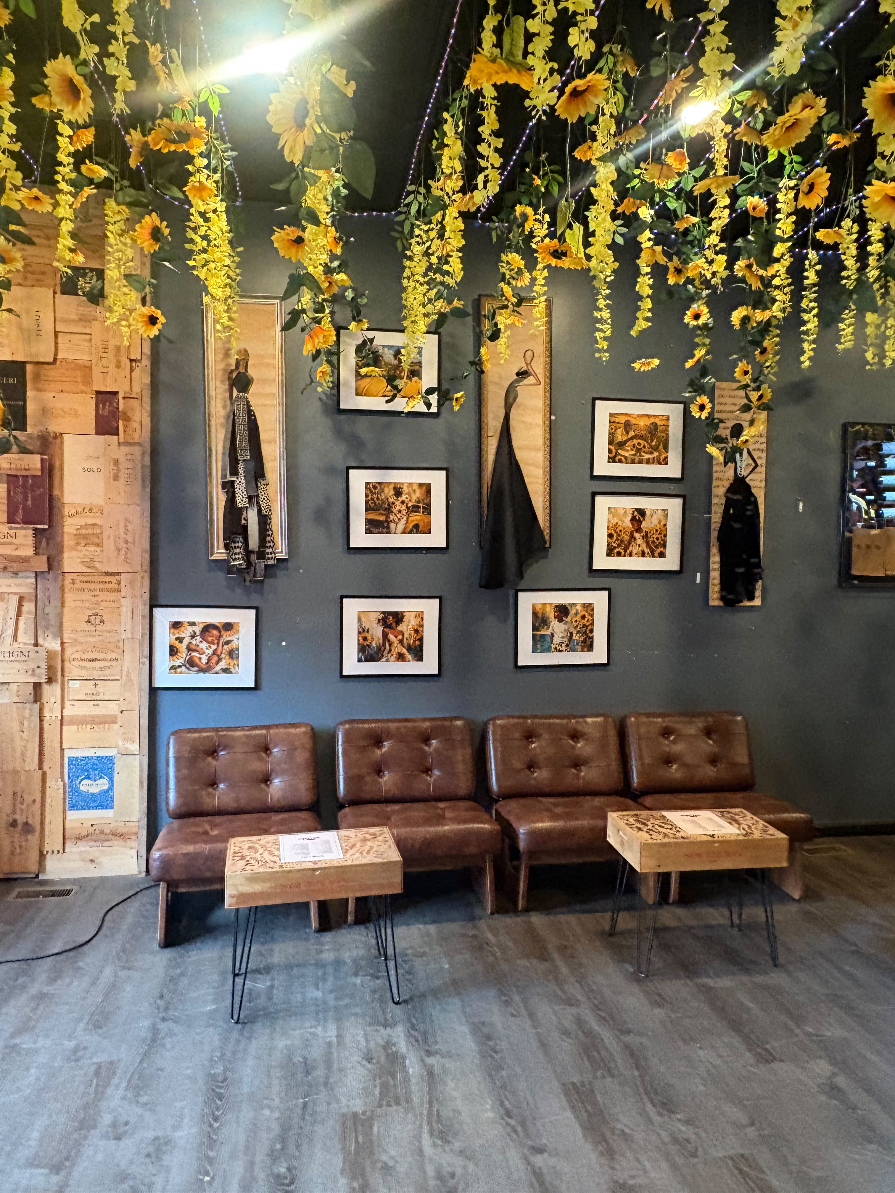 Intertwine's gallery wall — framed art and textile figures beneath a canopy of hanging sunflowers, above a row of leather lounge chairs